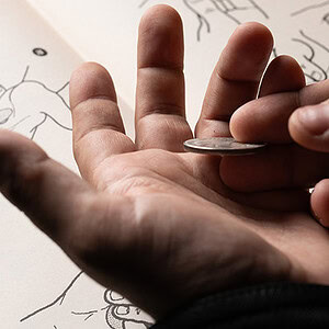 A person balancing a coin on their fingers over a sketchbook with drawings.