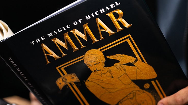 A magician performing a card trick with a black and gold book titled "The Magic of Michael Ammar".
