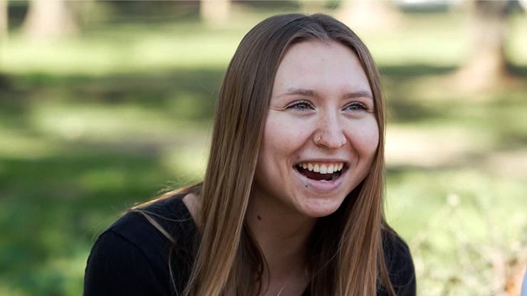 A smiling young woman enjoying a magic show outdoors in a park setting.