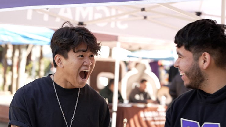 A lively outdoor event with two young men engaging in a joyful conversation under a tent.