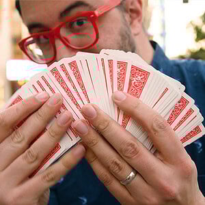 A man with red glasses holding a fan of playing cards outdoors.