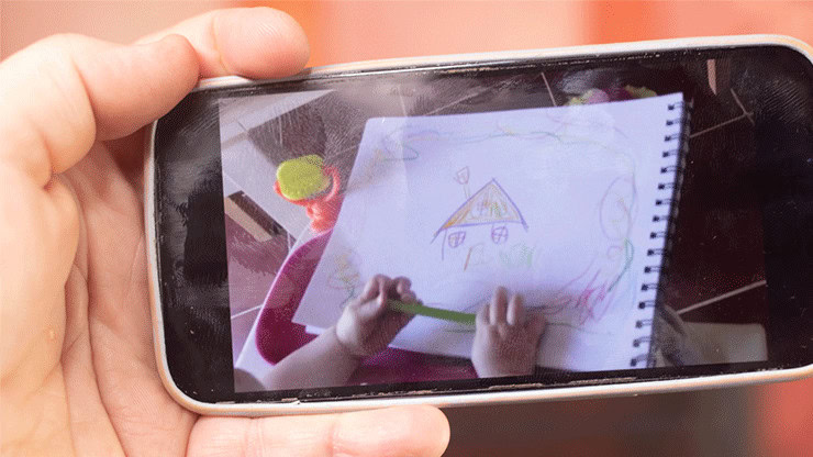 Child drawing a colorful house on paper with a green crayon.