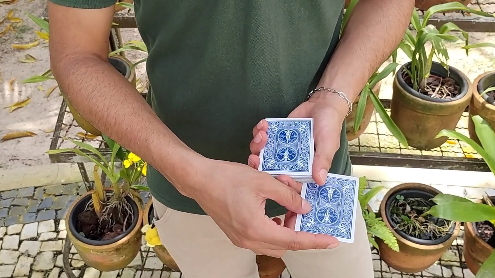 Close-up of a person performing card magic outdoors with potted plants in the background.