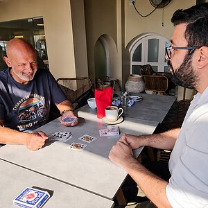 Nikolas Mavresis performing No Explanation at a cafe table — two men with playing cards