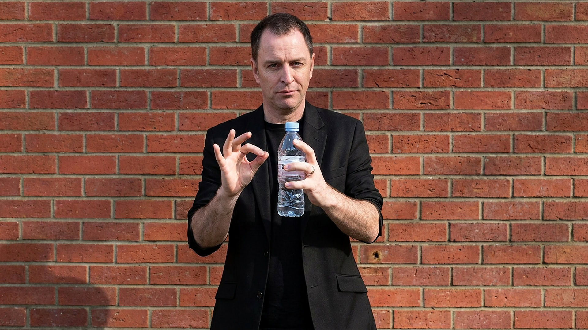 reKap by Richard Griffin magic trick - performer demonstrating the bottle cap effect with a water bottle against a brick wall